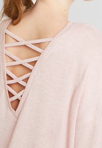 Jasnoróżowy pleciony top z głębokim dekoltem w serek z krzyżującymi się detalami. Miękka tekstura, luźny krój i opadające ramiona.