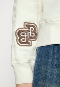 Manche de sweat-shirt blanc ornée d'un logo brodé marron texturé avec un design double 'B'. Porté avec un jean denim bleu.