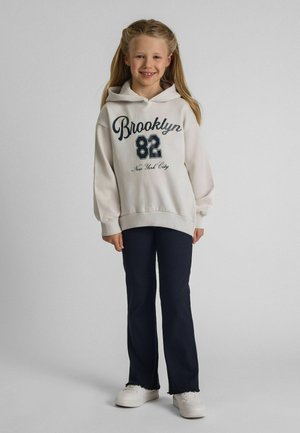 Uśmiechnięta dziewczyna z długimi blond włosami, ubrana w biały kaptur "Brooklyn 82 New York City", czarne spodnie i białe trampki, stojąca na szarym tle.