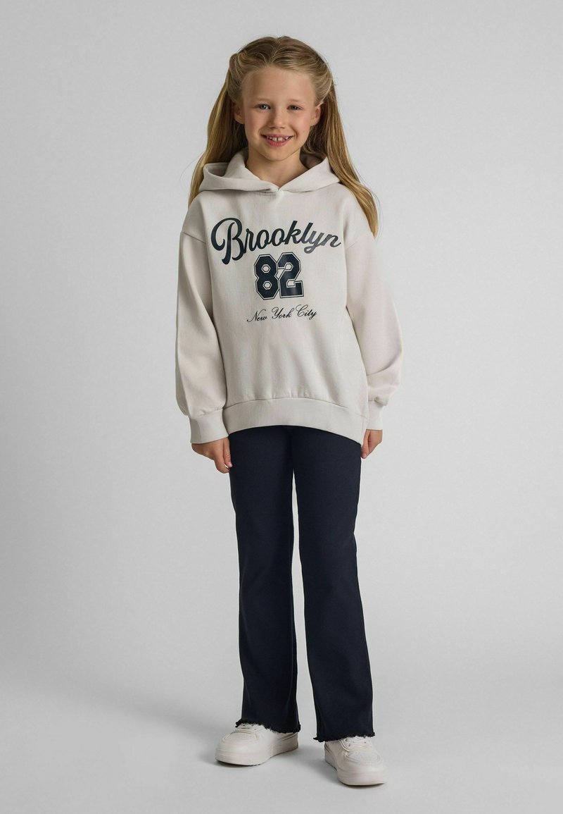 Lächelndes Mädchen mit langen blonden Haaren, das einen weißen Hoodie mit der Aufschrift "Brooklyn 82 New York City", schwarze Hose und weiße Turnschuhe trägt und vor einem grauen Hintergrund steht.