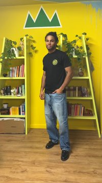 T-shirt preta com logótipo verde, jeans azuis e sapatos pretos. Parede amarelo vivo com design de montanha verde; estantes com livros e plantas.