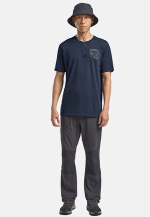 Uomo in piedi che indossa un cappello da pescatore nero, una t-shirt a maniche corte blu navy con grafica, pantaloni da trekking grigio scuro e scarpe da trekking nere.