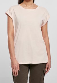 Kobieta ubrana w jasnoróżową koszulę z krótkim rękawem z podwiniętymi mankietami oraz ciemnozielone spodnie cargo, stojąca na jednolitym tle.