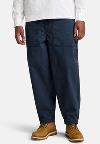 Pantaloni ampi di colore blu scuro in cotone, con grandi tasche a toppa e una vestibilità rilassata, abbinati a stivali marroni con lacci.