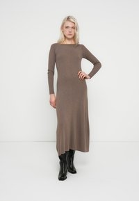 Vestido largo ajustado de punto marrón con mangas largas y escote cuadrado. Se combina con botas negras hasta la rodilla. Textura suave y diseño minimalista.