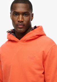 Sudadera de color coral con un bolsillo frontal, capucha con cordones y logotipo bordado. Cuello de cuadros en capas visible. Textura suave.