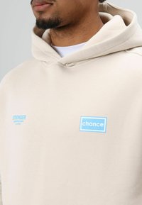 Beige hoodie met een grote voorkantzak, een capuchon met trekkoorden en blauwe bedrukte accenten "STRONGER" en "chance." Gladde stoftextuur.