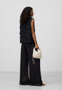 Top nero senza maniche con dettagli in frange testurizzate, abbinato a pantaloni neri larghi e una borsa crema con manico curvo.