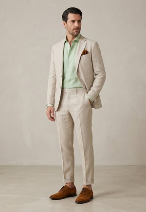 Mann trägt einen beigen Anzug mit einem hellgrünen Hemd, braunem Einstecktuch und braunen Schnürschuhen, steht mit einer Hand in der Tasche.