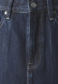 Jeans de denim oscuro con cintura alta, que presentan un botón frontal, cremallera y costuras amarillas contrastantes a lo largo de las costuras y los bolsillos.