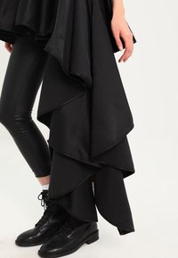 Schwarzes, geschichtetes Kleid mit einem fließenden, asymmetrischen Design. Kombiniert mit schwarzen Lederhosen und Stiefeln mit Knöchelschaft. Glatte Textur, modernes Silhouette.