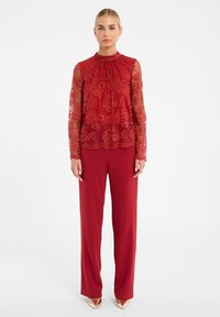 Rode kant blouse met lange mouwen en hoge hals, gecombineerd met losse rode broeken en gouden puntige hakken.