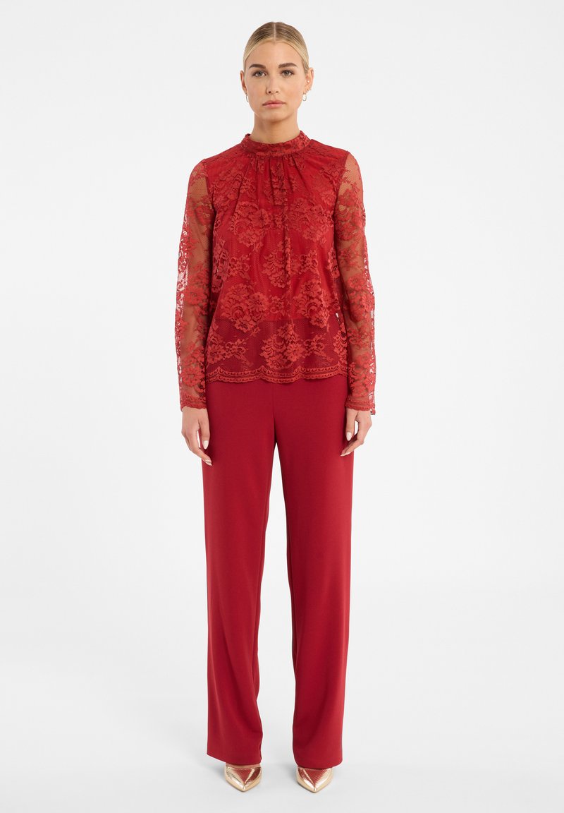 Rode kant blouse met lange mouwen en hoge hals, gecombineerd met losse rode broeken en gouden puntige hakken.