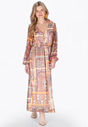 Kobieta o długich blond włosach, ubrana w długą, wzorzystą sukienkę w ciepłych tonach i beżowe botki, uśmiechająca się, nosząca pomarańczowe, wyróżniające się kolczyki.