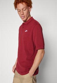 Μπορντό πόλο μπλούζα από υφασμάτινη υφή, με λευκό λογότυπο Nike στο αριστερό στήθος. Κοντά μανίκια με κλασικό γιακά.