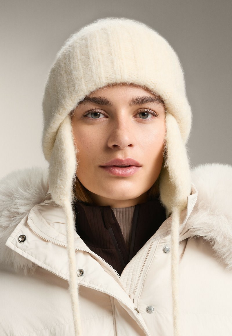 Biela pletená čiapka s chráničmi na uši, vyrobená z mäkkého materiálu. Nosená s béžovou prešívanou bundou s kapucňou lemovanou kožušinou a tmavou vnútornou vrstvou.