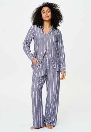 Femme aux cheveux bouclés portant un pyjama ample rayé verticalement bleu et blanc avec des boutons, debout pieds nus, une main dans la poche.