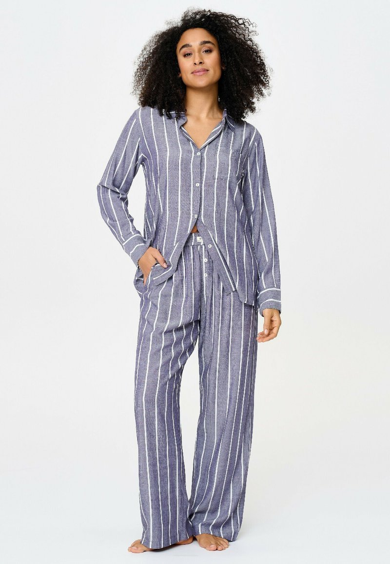 Femme aux cheveux bouclés portant un pyjama ample rayé verticalement bleu et blanc avec des boutons, debout pieds nus, une main dans la poche.