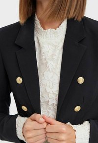 Černý dvojřadý blazer se zlatými knoflíky, s klopovým límcem a strukturovanými rameny, vrstvený přes bílý krajkový top.