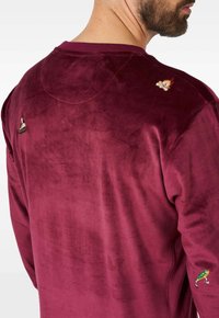 Bordo pamučna trenirka s baršunastom površinom i izvezenim detaljima na ramenima. Mekani materijal i okrugli izrez. Prikazan stražnji dio.