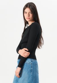 Mlada žena s dugom tamnom kosom koja nosi crnu majicu s dugim rukavima i plavu traper suknju, stoji naslonjena na bijelu pozadinu, s rukama prekriženim preko tijela.
