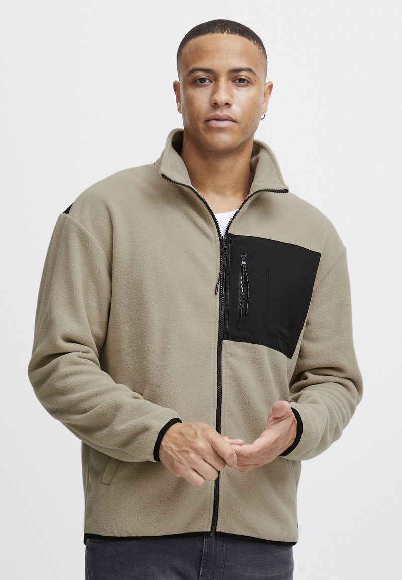 Chaqueta de forro polar de color marrón claro con un bolsillo en el pecho negro con cremallera, cuello alto y puños negros, que presenta una textura suave y cremallera de longitud completa.