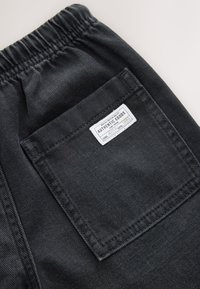 Pantalon en denim noir avec une ceinture élastique et une poche arrière ornée d'une étiquette blanche portant le texte « AUTHENTIC GOODS » et des détails.