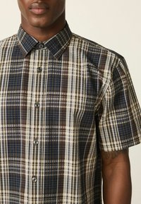 Károvaná košeľa s krátkym rukávom v odtieňoch hnedej, námorníckej modrej a krémovej farby. Má golier na zapínanie na gombíky a kontrastné gombíky. Textúrovaná látka.