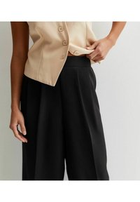 Beige knapp-up topp med v-ringning och korta ärmar, ihopparat med högmidjade svarta plisserade byxor, som visar släta tygtexturer.