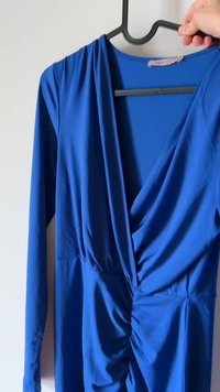 Abito blu a maniche lunghe in tessuto morbido, con una parte anteriore drappeggiata e dettagli arricciati in vita. Appeso a una gruccia nera su uno sfondo chiaro.