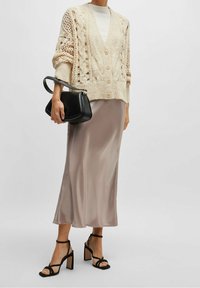 Kvinna som bär en krämfärgad stickad cardigan, vit topp, taupefärgad satin midi-kjol, svarta klacksandaler och håller en svart läderhandväska vid sidan.