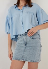 Camisa corta azul claro con mangas abullonadas y cierre de botones, combinada con una falda mini de mezclilla clara ajustada. Tejido suave y diseño casual.