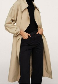 Manteau beige avec un large col, des manches longues et une coupe décontractée, associé à un pantalon noir. Tissu lisse avec peu de détails.