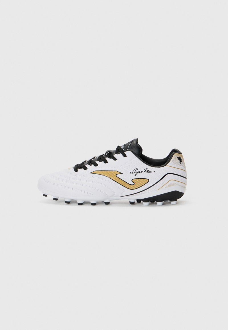 Chaussure de soccer athlétique blanche avec un dessus texturé, des accents noirs et un logo doré. Elle dispose d'une semelle à crampons pour une meilleure adhérence sur les surfaces en herbe.