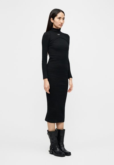 Μαύρο φόρεμα με μακριά μανίκια, ψηλό λαιμό και εφαρμοστή γραμμή, κατασκευασμένο από ελαστικό ύφασμα. Συνδυάζεται με μαύρες δερμάτινες μπότες μέχρι το γόνατο με αγκράφες.