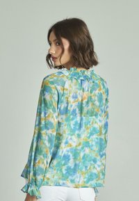 Leichte, blau-grüne Batikbluse mit rüschigem Ausschnitt, langen Ärmeln und elastischen Bündchen. Verfügt über eine lockere Passform und fließenden Stoff.