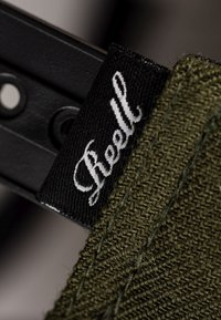 Etiqueta verde de tela tejida con letras blancas "Reell", unida a un objeto metálico negro con agujeros circulares y un borde plano.