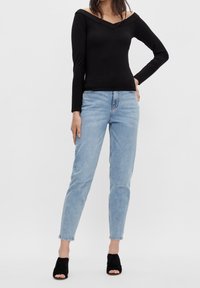 Eine Frau steht da und trägt ein schwarzes, schulterfreies Langarmoberteil, hellblaue, hoch taillierte Jeans und schwarze, offene High Heels.