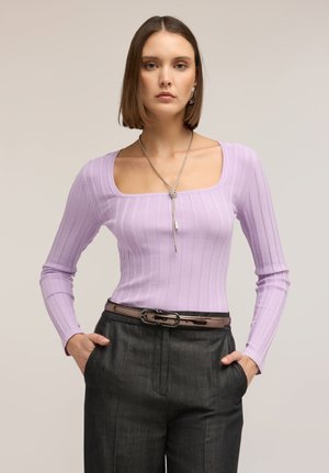 Mujer con cabello castaño liso que lleva una blusa de manga larga color lila con textura acanalada y escote cuadrado, pantalones gris oscuro, cinturón marrón y collar plateado.