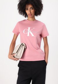 Lys rosa bomull T-skjorte med korte ermer, som har et hvitt "CK"-logo og "Calvin Klein Jeans"-tekst. Modellen holder en beige håndveske.