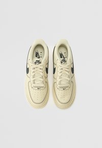 Sapatilhas Nike Air Force 1 em pele creme com detalhes em preto, apresentando um biqueira perfurada, atacadores planos e uma sola de borracha.