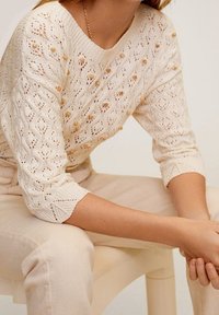 Szydełkowy sweter w kolorze kremowym z diamentowym wzorem, teksturowany małymi bąbelkami. Posiada szeroki dekolt i rękawy o długości trzech ćwiartek.