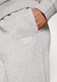 Sivé tepláky vyrobené z mäkkej tkaniny, s vyšitým logom Reebok v bielej farbe na prednej kapsičke a rebrovanými manžetami pre pohodlné nosenie.