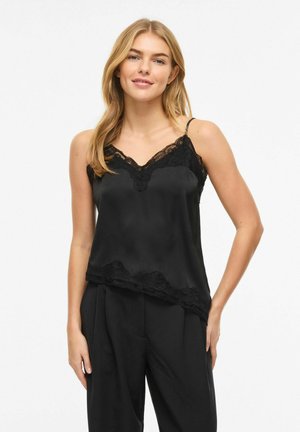 Blondína v čiernom saténovom tielku s čipkovaným lemom a čiernych nohaviciach, stojaca s jednou rukou v vrecku na bielom pozadí.