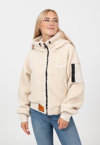 Kremowa polarowa kurtka z czarnym zamkiem błyskawicznym, oversize'owy kaptur, elastyczne mankiety i boczna kieszeń. Posiada teksturowany materiał i pomarańczową metkę.