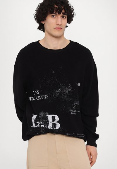 Pull noir surdimensionné avec texte graphique blanc et motifs de éclaboussures. Texture côtière et larges poignets. Associé à un pantalon beige.