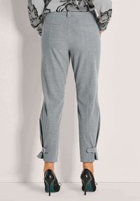Vrouw in een grijze, op maat gemaakte broek met enkelgespen en zwarte hoge hakken, staand tegen een effen lichte achtergrond.