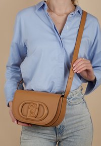 Bolso de cuero marrón claro cruzado con logotipo en relieve, forma rectangular, diseño plano y correa ajustable, combinado con una camisa azul claro y vaqueros.