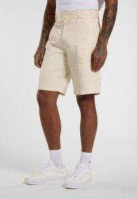 Beige Shorts mit einem maßgeschneiderten Schnitt, flacher Vorderseite, Gürtelschlaufen und Seitentaschen, kombiniert mit weißen Sneakers und Knöchelsocken.