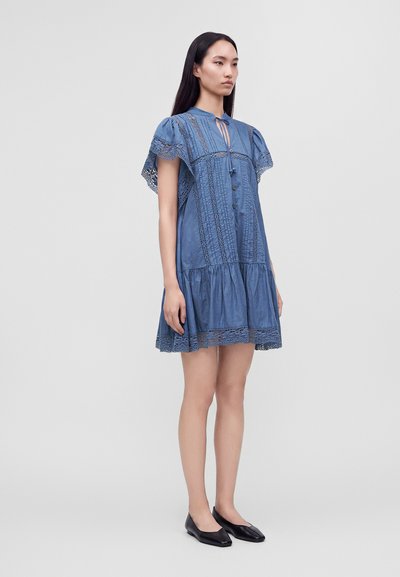Femme debout portant une robe bleue à manches courtes avec détails en dentelle et boutons, associée à des chaussures plates noires sur un fond clair uni.